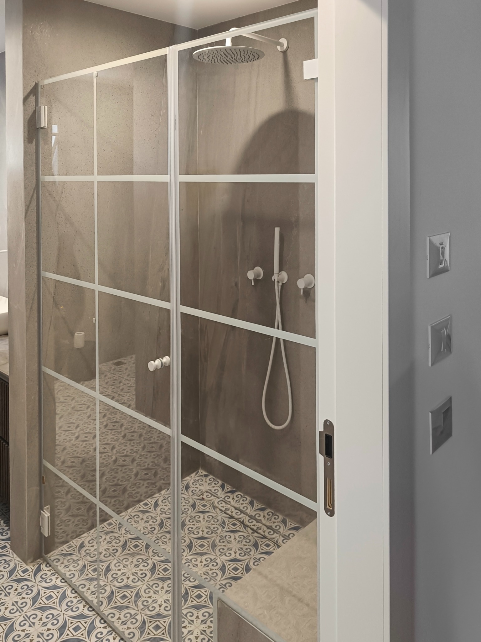 Nowoczesna kabina prysznicowa z transparentnymi szklanymi drzwiami w białej ramie z podziałami, widoczny panel prysznicowy i wzorzysta podłoga.