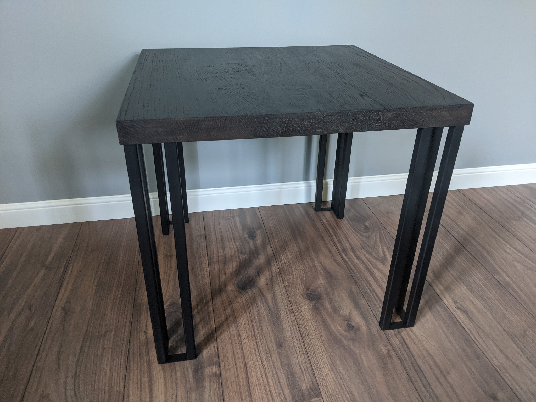 Kwadratowy stolik kawowy z blatem z ciemnego drewna i czarnymi, podwójnymi nogami z metalu, ustawiony na podłodze z paneli w odcieniach brązu.