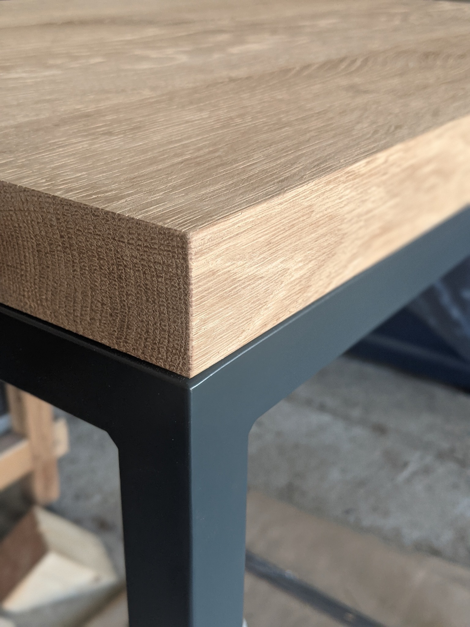 Fragment blatu stołu z jasnego drewna dębowego, wspartego na metalowej, grafitowej ramie o kwadratowym profilu, widoczny z bliska, z surowym betonowym tłem.