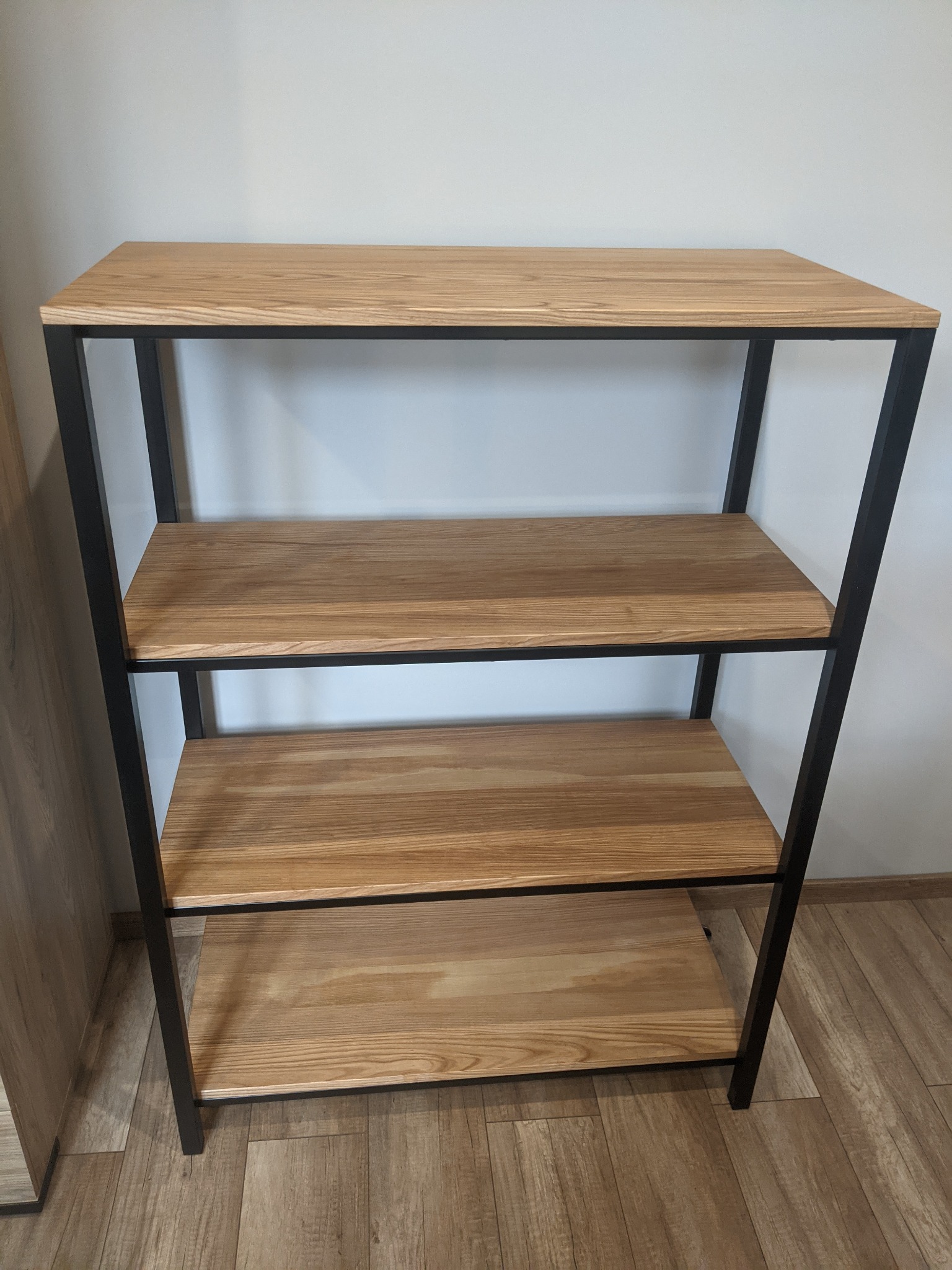 Regał z trzema półkami z jasnego drewna, osadzony na czarnej, metalowej konstrukcji, stojący na podłodze imitującej drewno, z widoczną białą ścianą w tle.