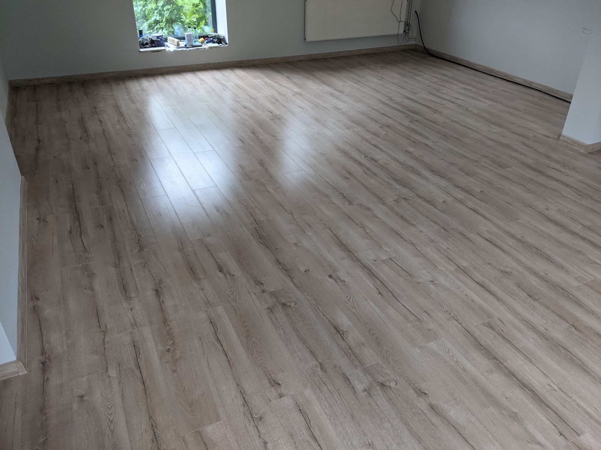 Jasne, ułożone panele podłogowe z widocznym wzorem drewna, świeżo po montażu w pustym pokoju z oknem i grzejnikiem.