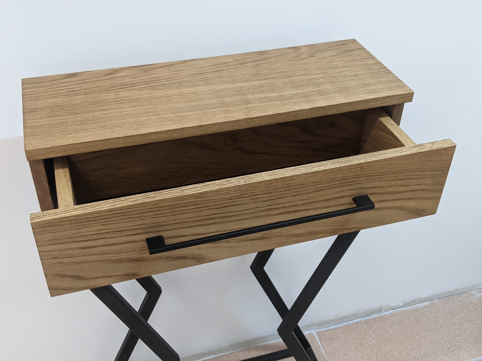Drewniana szafka nocna z wysuniętą szufladą, wsparta na czarnym metalowym stelażu w kształcie litery X, na tle białej ściany i beżowej podłogi.
