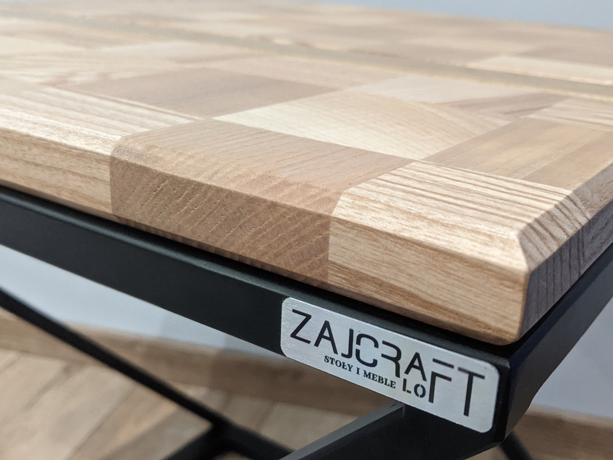 Blat stołu z drewna klejonego w szachownicę, osadzony na metalowym, czarnym stelażu z widocznym logo 'ZAJCRAFT STOŁY I MEBLE LO.'