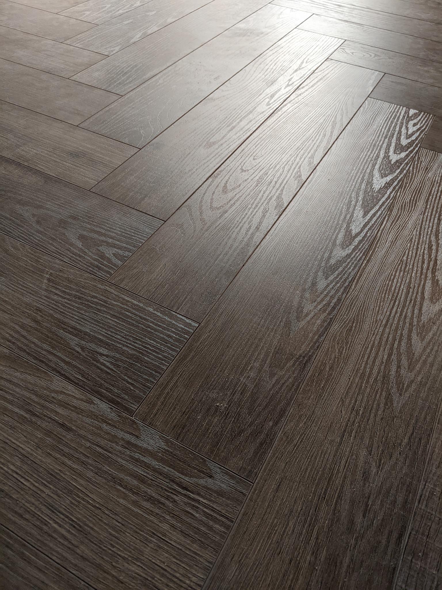 Ułożone panele podłogowe w jodełkę, z widoczną strukturą drewna i delikatnym połyskiem, oświetlone z góry, tworzące geometryczny wzór.