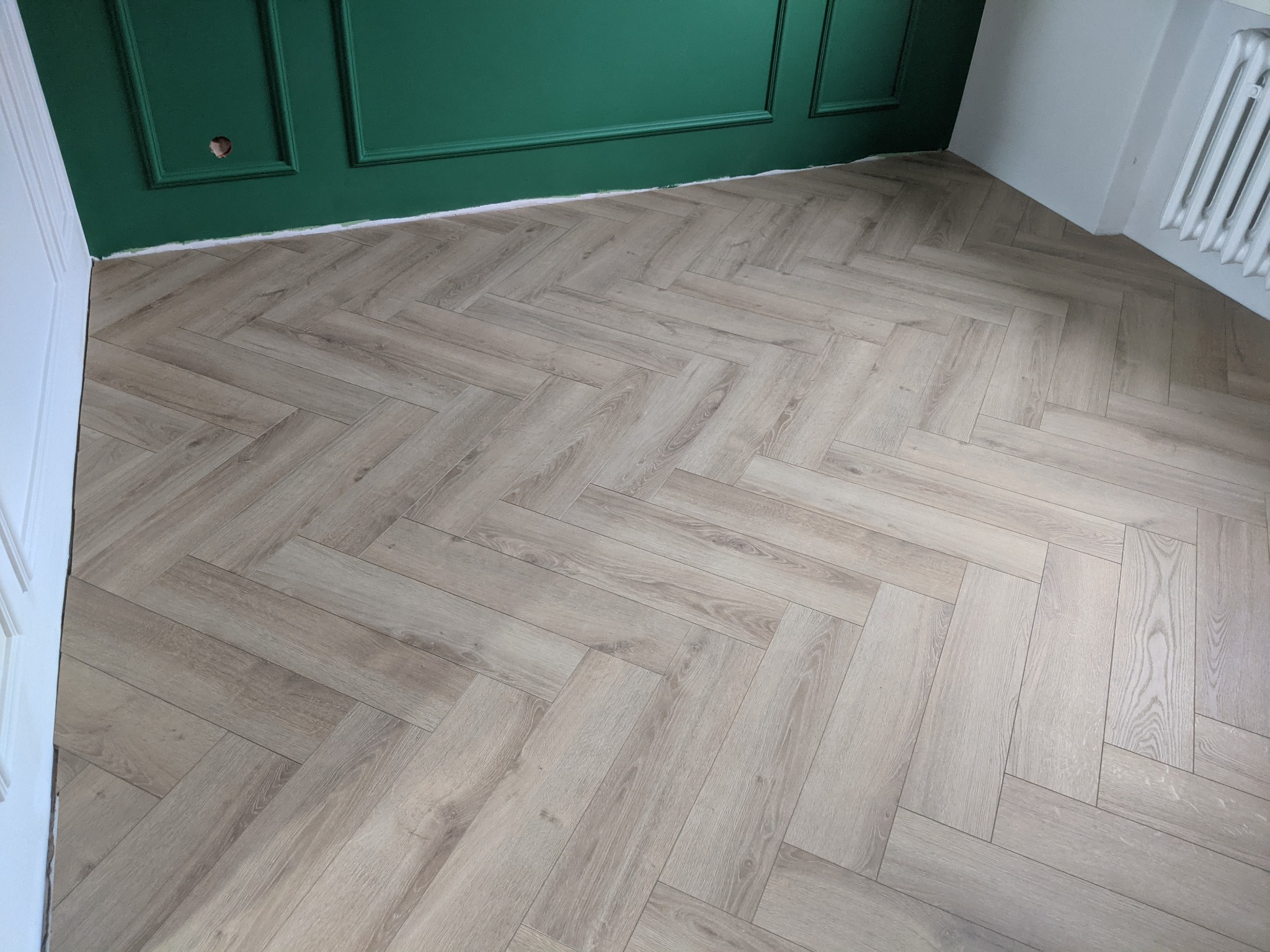 Ułożona podłoga z paneli w jodełkę, jasne drewno, zielona ściana z dekoracyjnymi listwami, białe listwy przypodłogowe i biały grzejnik.