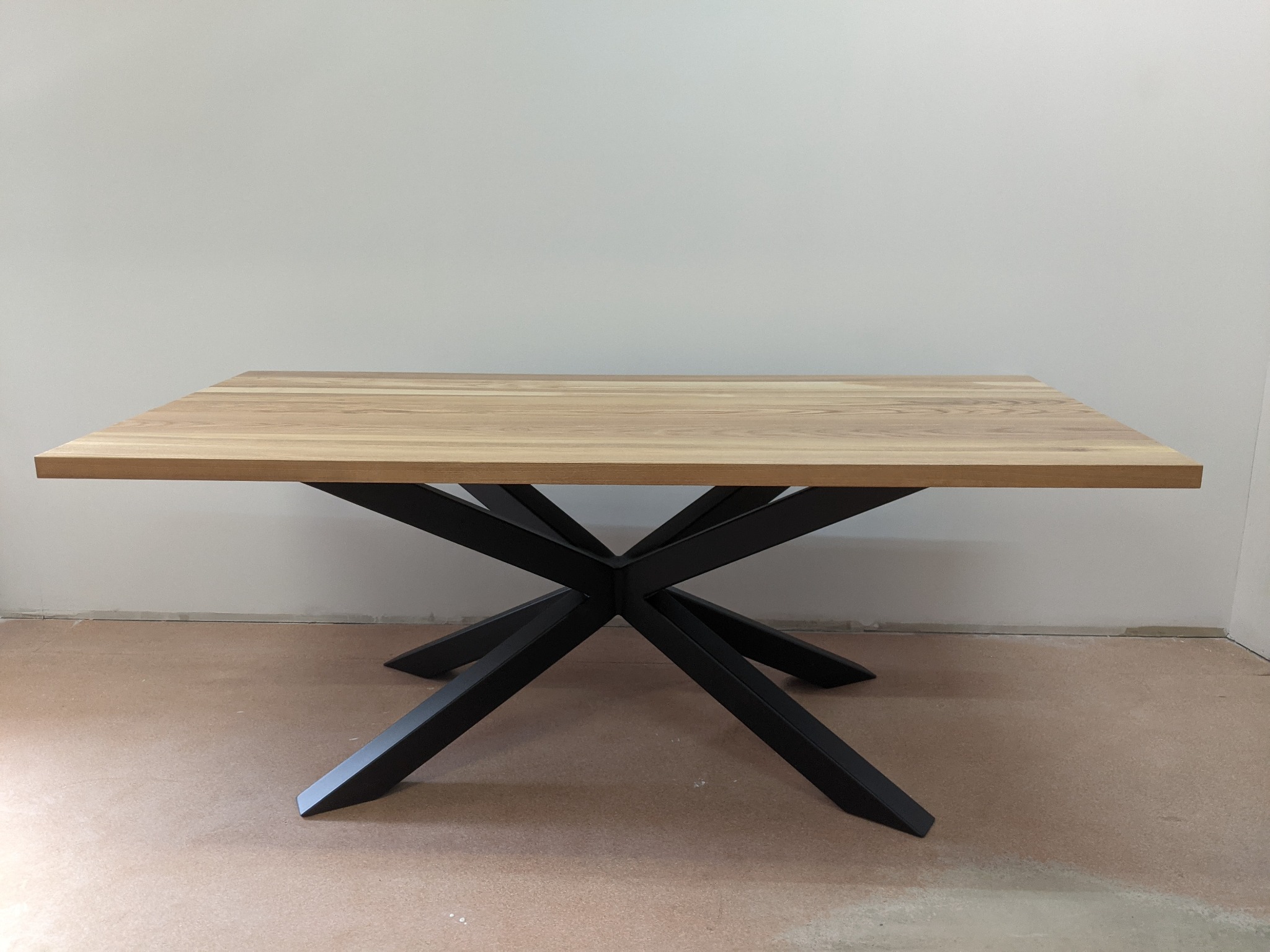 Kwadratowy stół z jasnego drewna z czarną, metalową podstawą w kształcie gwiazdy, na tle białej ściany i surowej podłogi.