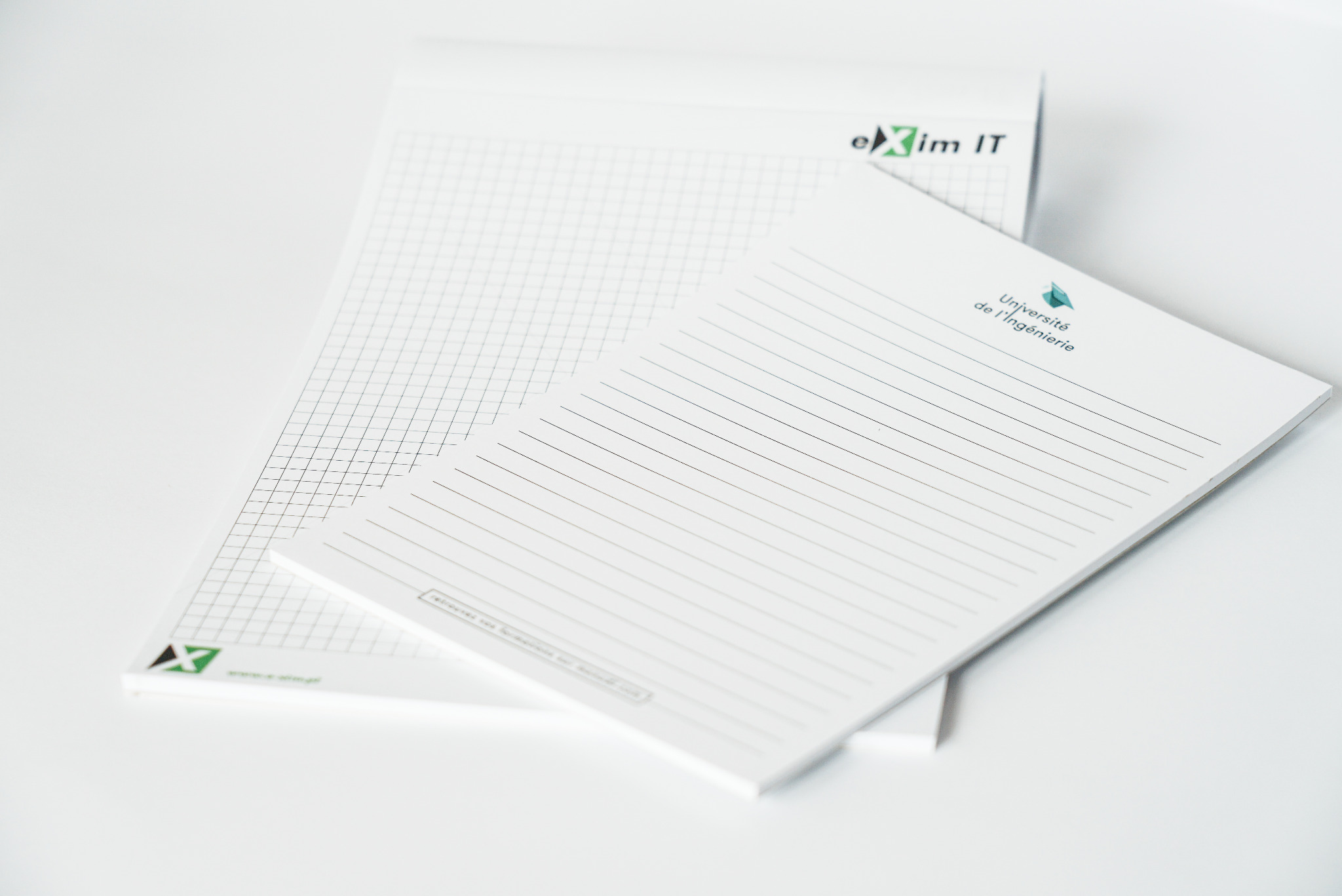 Dwa notesy firmowe leżące na białym tle. Jeden w kratkę z logo 'eXim IT', drugi w linie z logo 'Université de l'Ingénierie'.