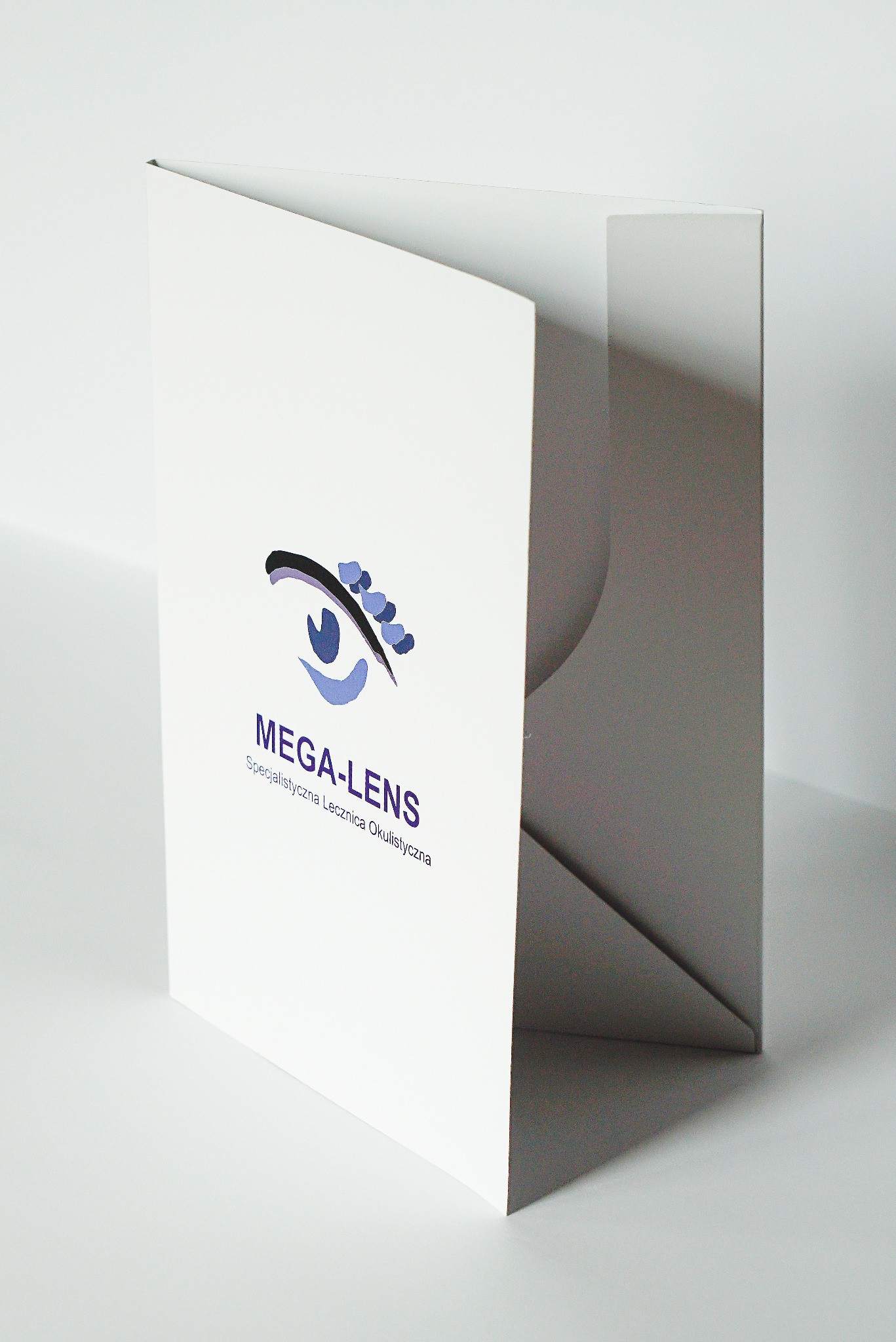Biała, otwarta teczka firmowa z logo Mega-Lens, specjalistycznej lecznicy okulistycznej, na jasnym tle.
