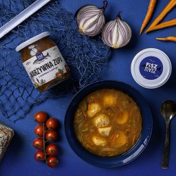 Bartosz Dymek - Kompozycja flat lay na niebieskim tle: słoik i pokrywka z logo marki zupy warzywnej, talerz z zupą z pulpetami, cebula przekrojona na pół, gałązka pomidorków cherry, marchewki, chleb i łyżka.