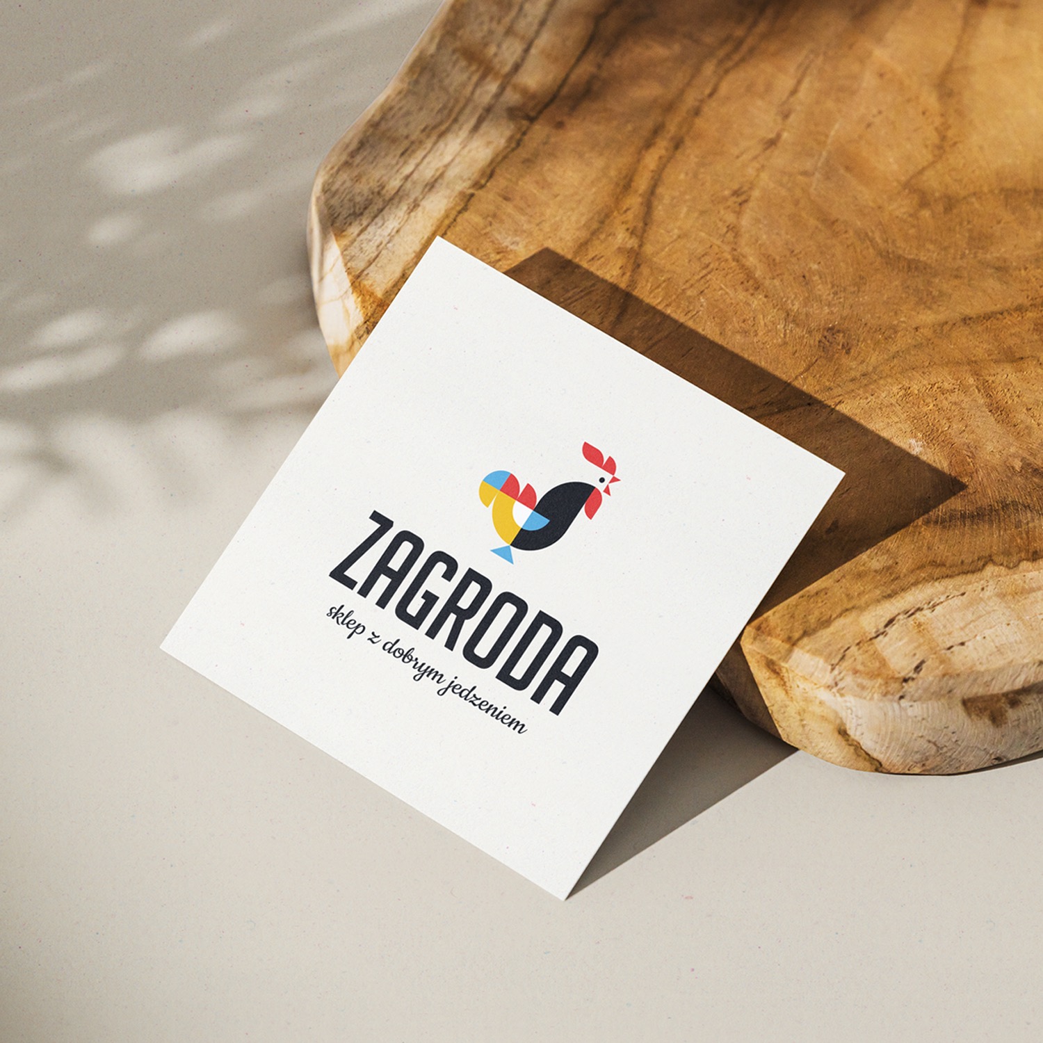Kwadratowa wizytówka z logo 'ZAGRODA sklep z dobrym jedzeniem' na drewnianej desce w jasnym oświetleniu.