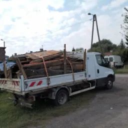 Biała ciężarówka z otwartą skrzynią ładunkową, wypełnioną pociętymi elementami drewnianymi i fragmentami dachu, zabezpieczonymi pomarańczowymi pasami transportowymi, zaparkowana na poboczu drogi...