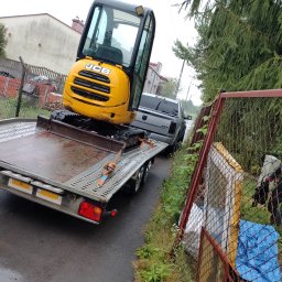 Clean Manager - Żółta mini koparka JCB na przyczepie transportowej, zabezpieczona pasami, ciągnięta przez srebrnego pickupa Dodge na mokrej, asfaltowej drodze w pobliżu ogrodzenia z siatki.