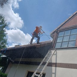 Clean Manager - Pracownik w uprzęży myje dach domu z drabiny, używając myjki ciśnieniowej. Widok z dołu na błękitne niebo z chmurami i fragment budynku.