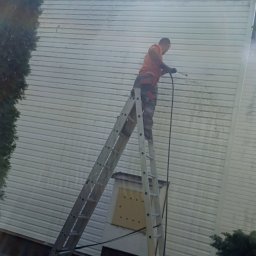 Clean Manager - Pracownik na drabinie czyści elewację sidingową myjką ciśnieniową. Widoczne zabrudzenia i strumień wody. Częściowo oświetlona elewacja w trakcie czyszczenia.