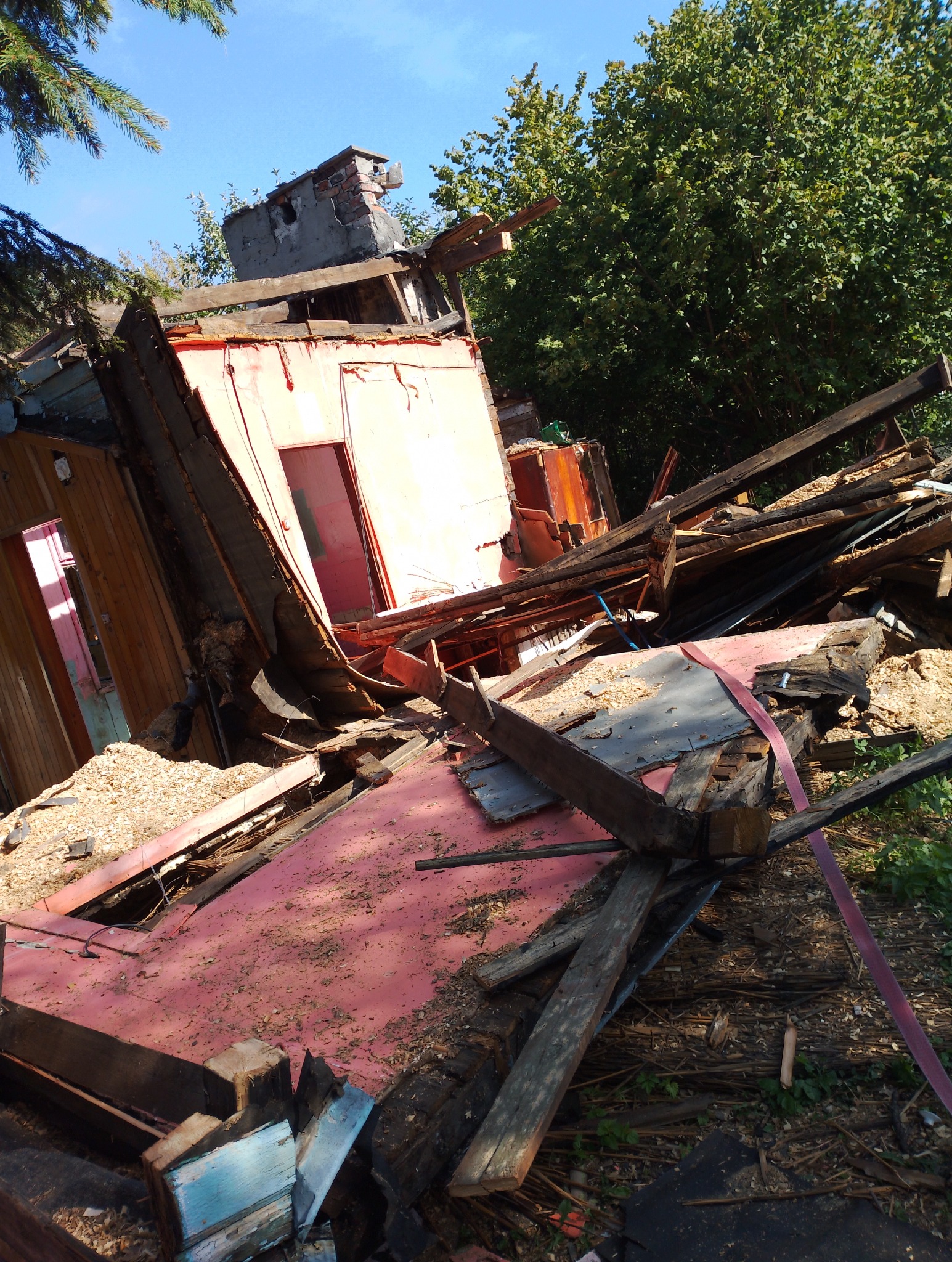 Zdjęcie przedstawia zniszczony, częściowo rozebrany budynek z elementami czerwonej elewacji i drewnianej konstrukcji, otoczony gruzem i zielenią, widziany z perspektywy z dołu.