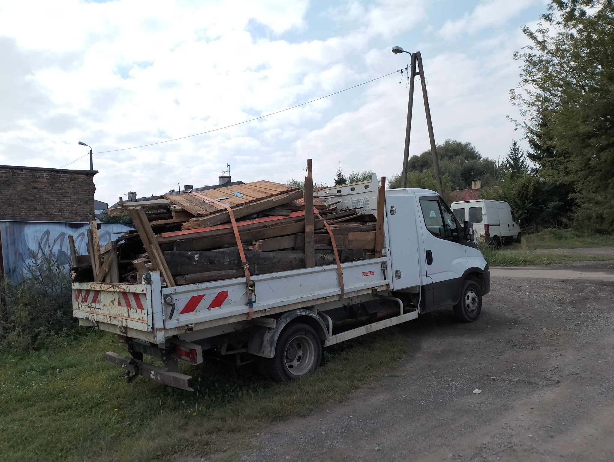 Biała ciężarówka z otwartą skrzynią ładunkową, wypełnioną pociętymi elementami drewnianymi i fragmentami dachu, zabezpieczonymi pomarańczowymi pasami transportowymi, zaparkowana na poboczu drogi...