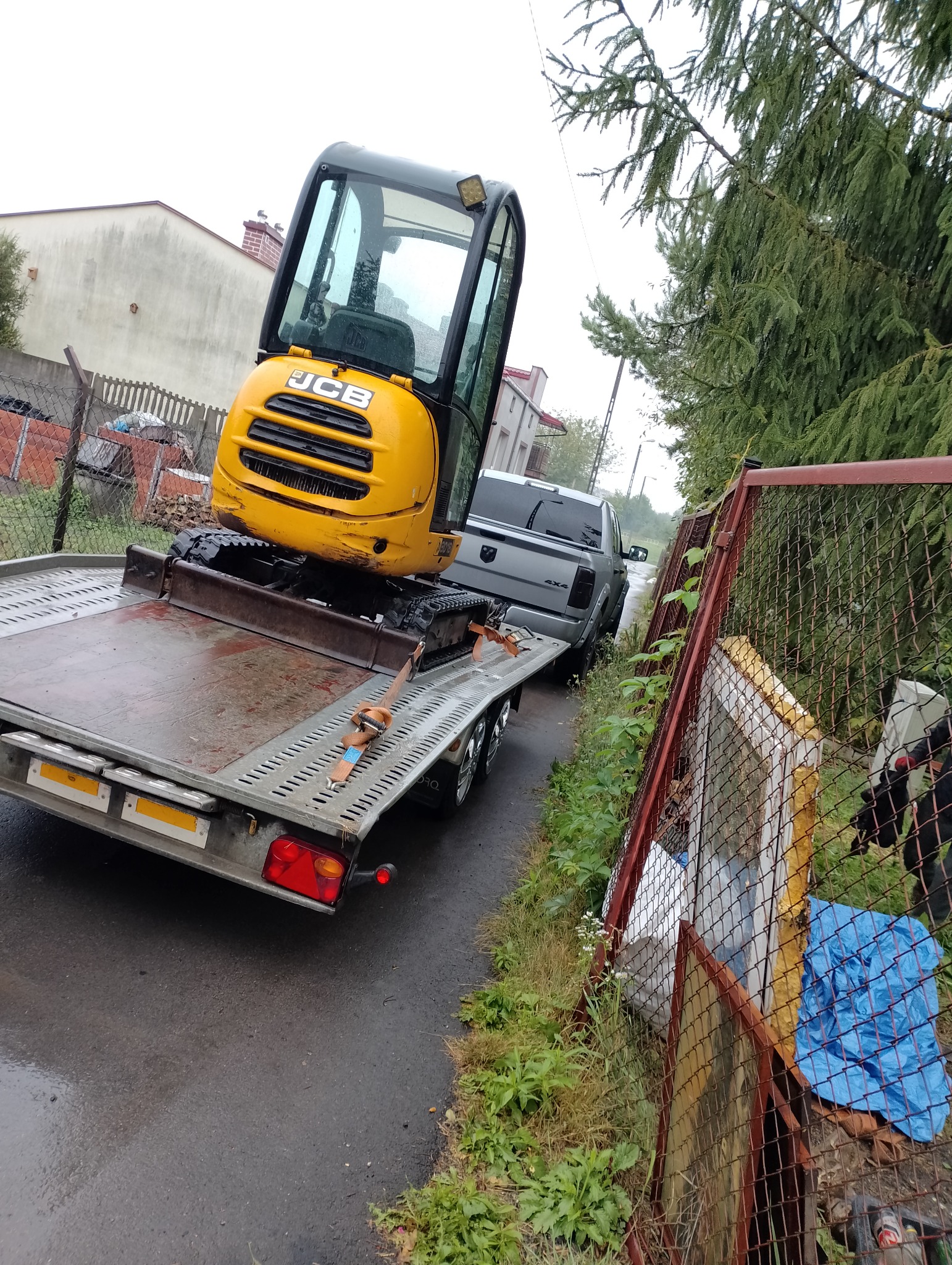 Żółta mini koparka JCB na przyczepie transportowej, zabezpieczona pasami, ciągnięta przez srebrnego pickupa Dodge na mokrej, asfaltowej drodze w pobliżu ogrodzenia z siatki.