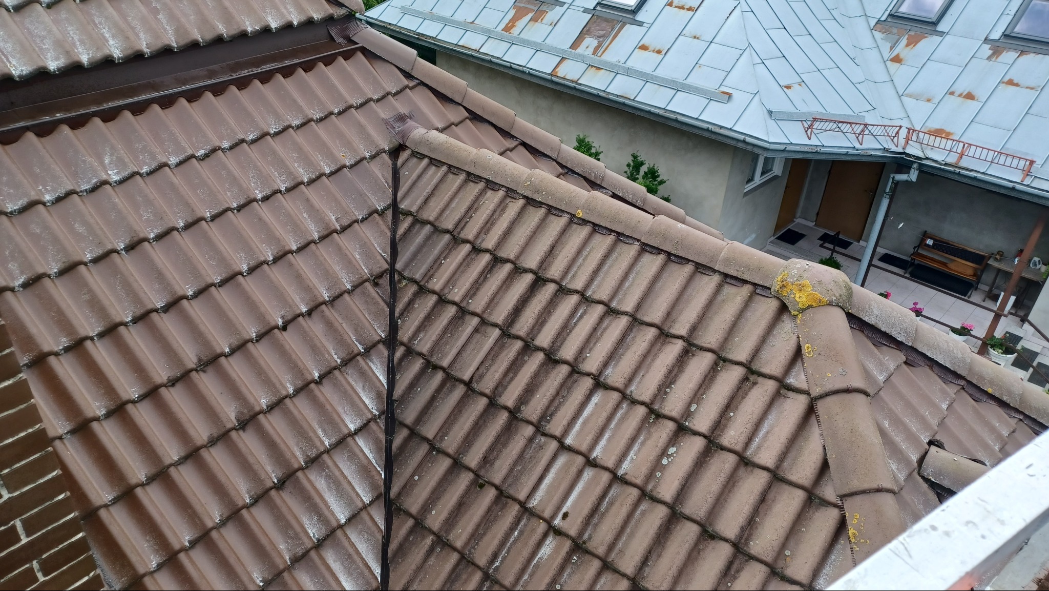 Ujęcie z góry na brązową dachówkę z widocznymi śladami zabrudzeń i mchu, sąsiedni budynek z szarą blachodachówką. Widoczne zacieki i nierównomierny koloryt.