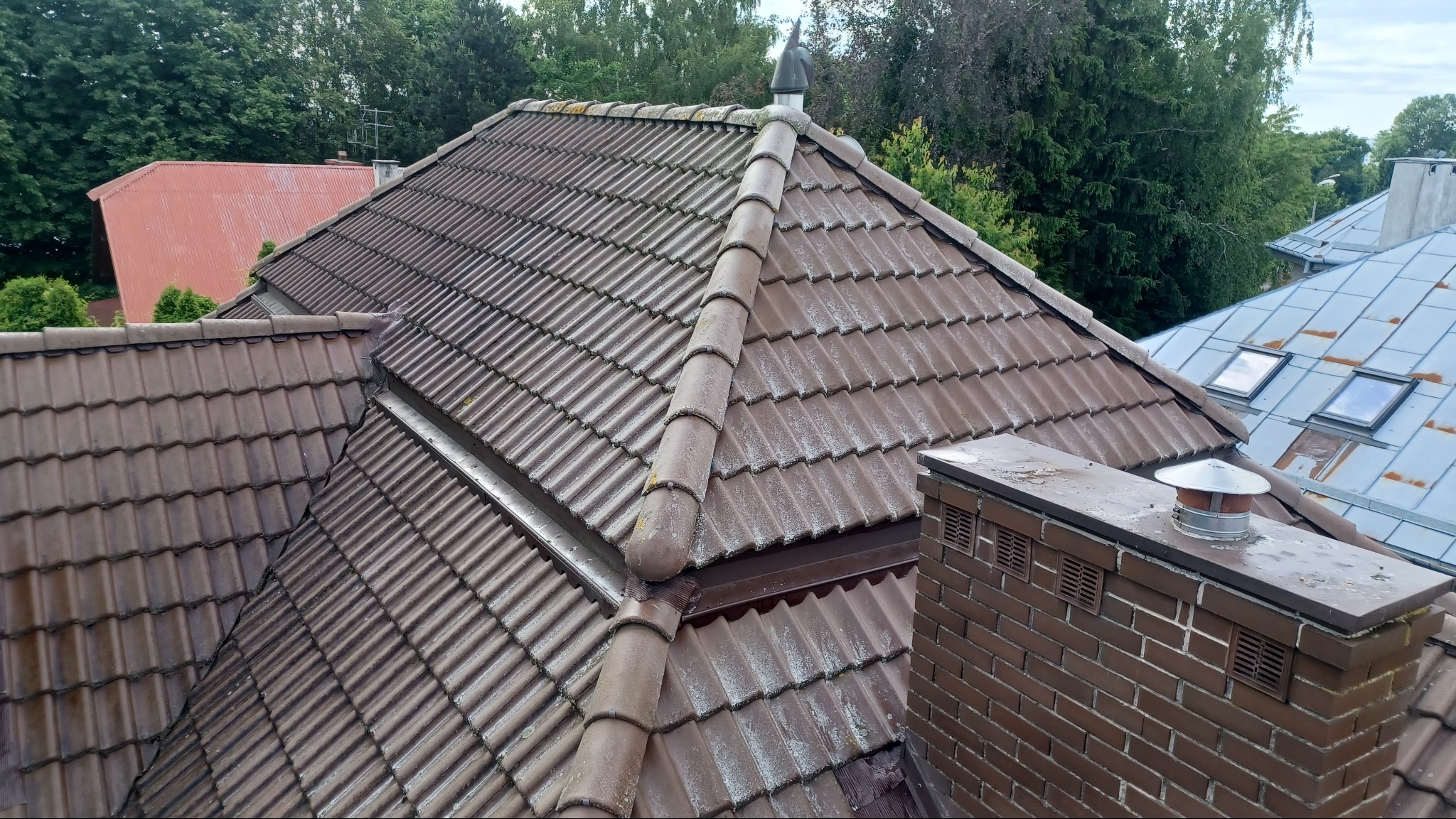 Brązowy dach z dachówki karpiówki z kominem z cegły, widoczny z góry, z zielonymi drzewami w tle i fragmentami innych budynków.