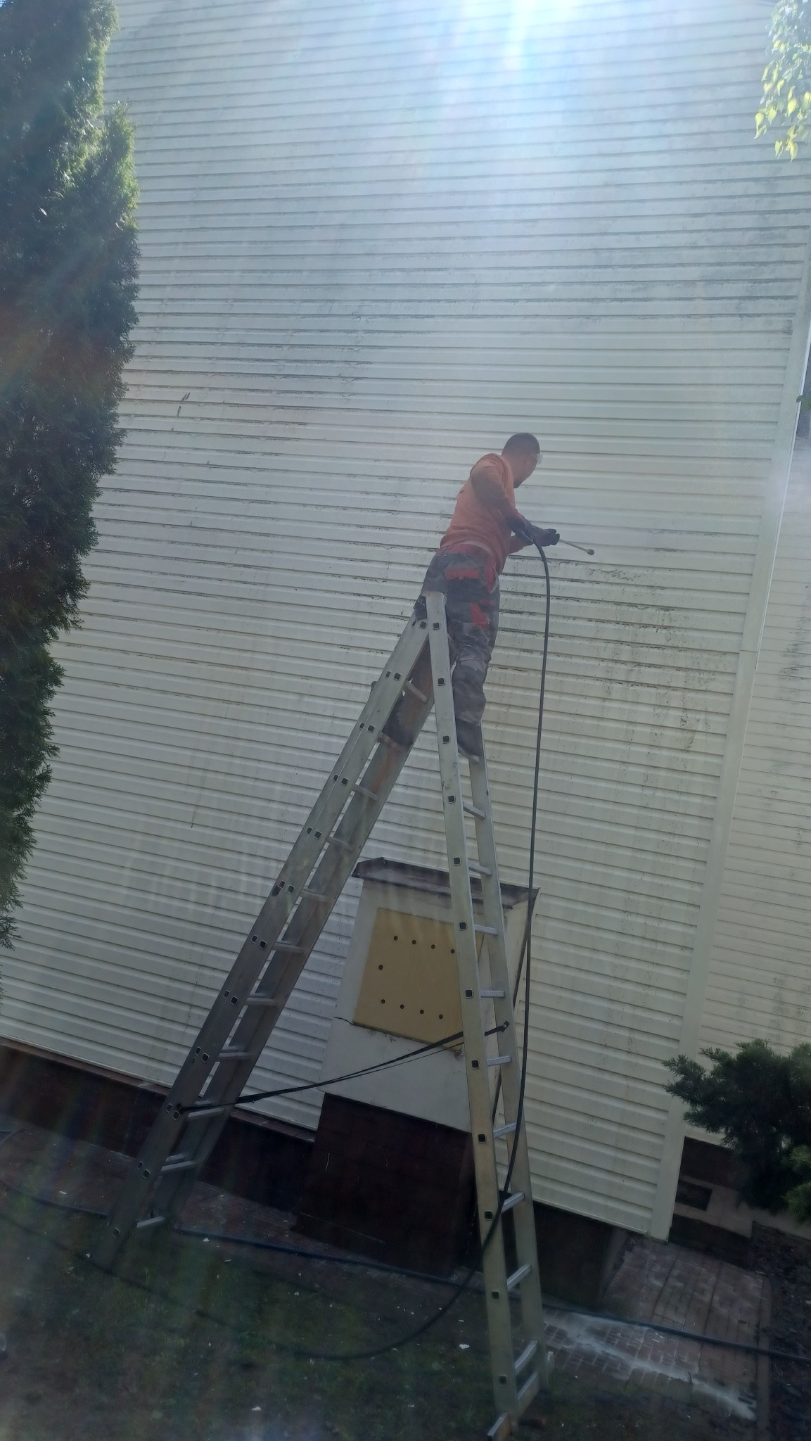 Pracownik na drabinie czyści elewację sidingową myjką ciśnieniową. Widoczne zabrudzenia i strumień wody. Częściowo oświetlona elewacja w trakcie czyszczenia.