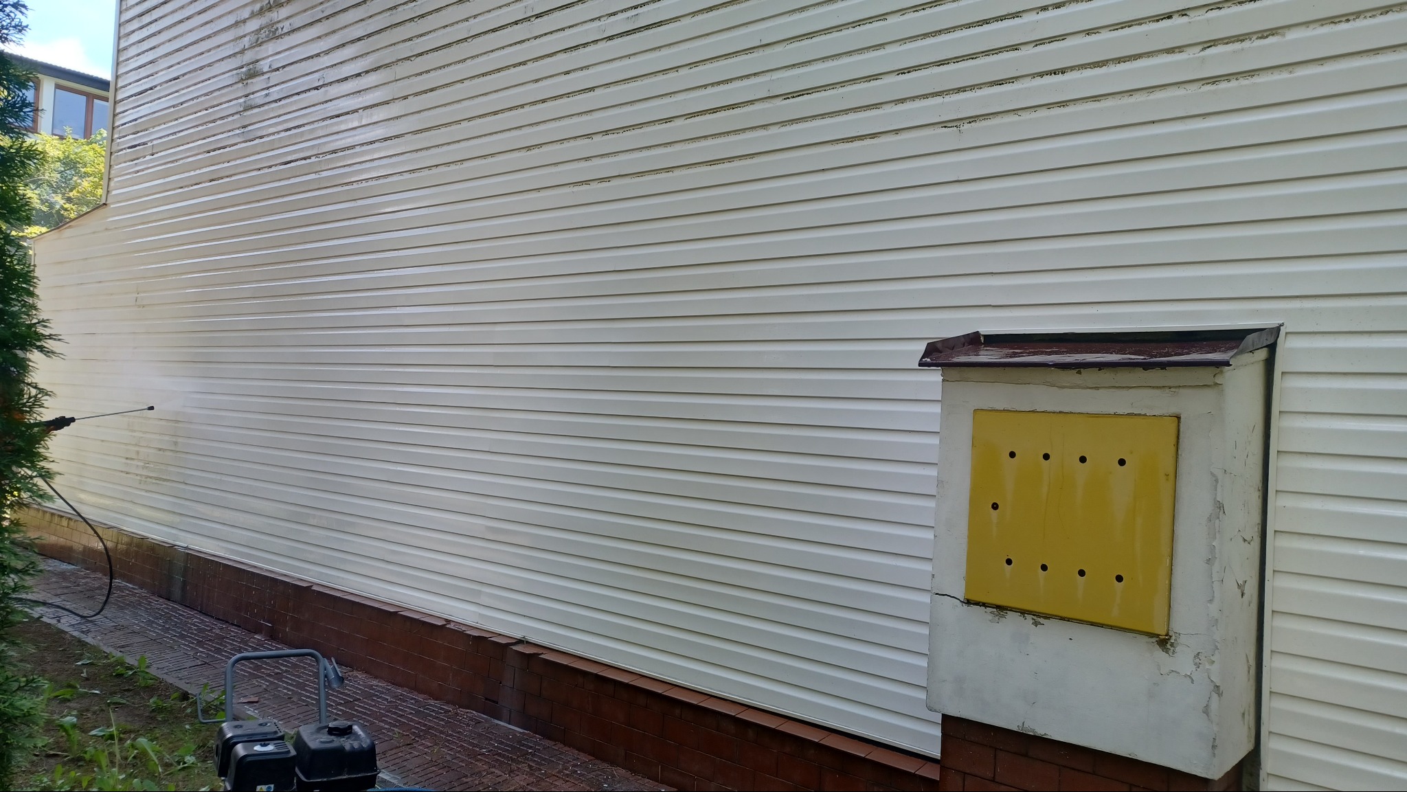 Czyszczenie elewacji sidingu myjką ciśnieniową. Widoczne zabrudzenia przed i po czyszczeniu, sprzęt do mycia. Ujęcie z dołu, z boku budynku.