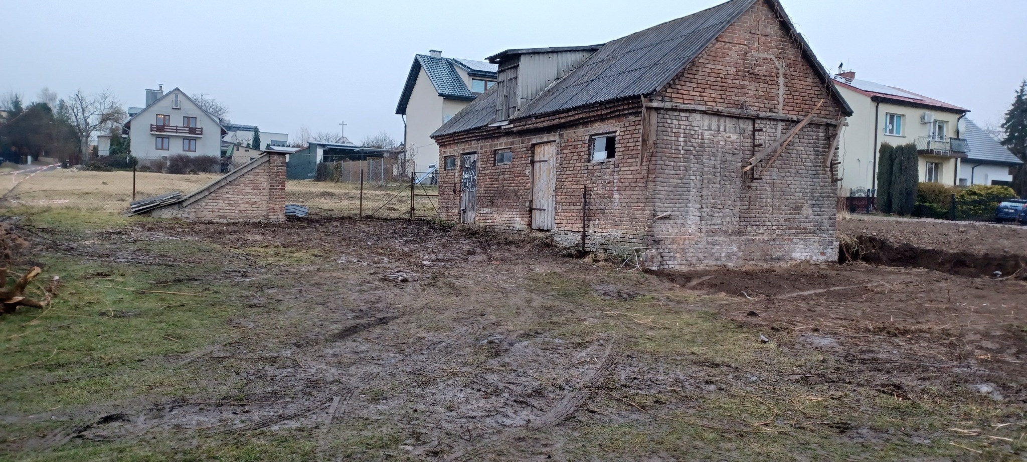 Stary, ceglany budynek gospodarczy z fragmentami rozbiórki i wykopami, na tle domów mieszkalnych. Teren błotnisty, ślady opon. Pochmurne niebo.