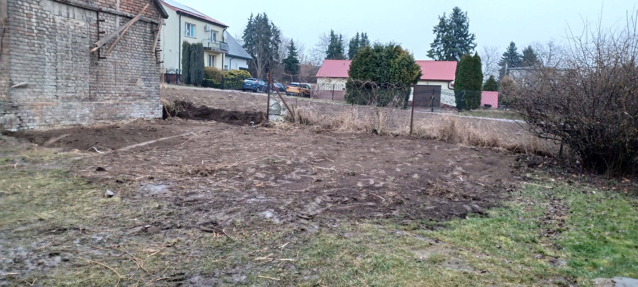 Przygotowany teren pod budowę, widoczna wykopana ziemia i wyrównany obszar. W tle budynki mieszkalne i roślinność. Prace ziemne w toku.