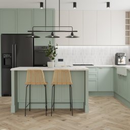 STUDIO OF3 - Nowoczesna kuchnia w odcieniach bieli i zieleni z drewnianą podłogą w jodełkę, wyspą z dwoma rattanowymi krzesłami barowymi oraz czarną lodówką z dystrybutorem wody.