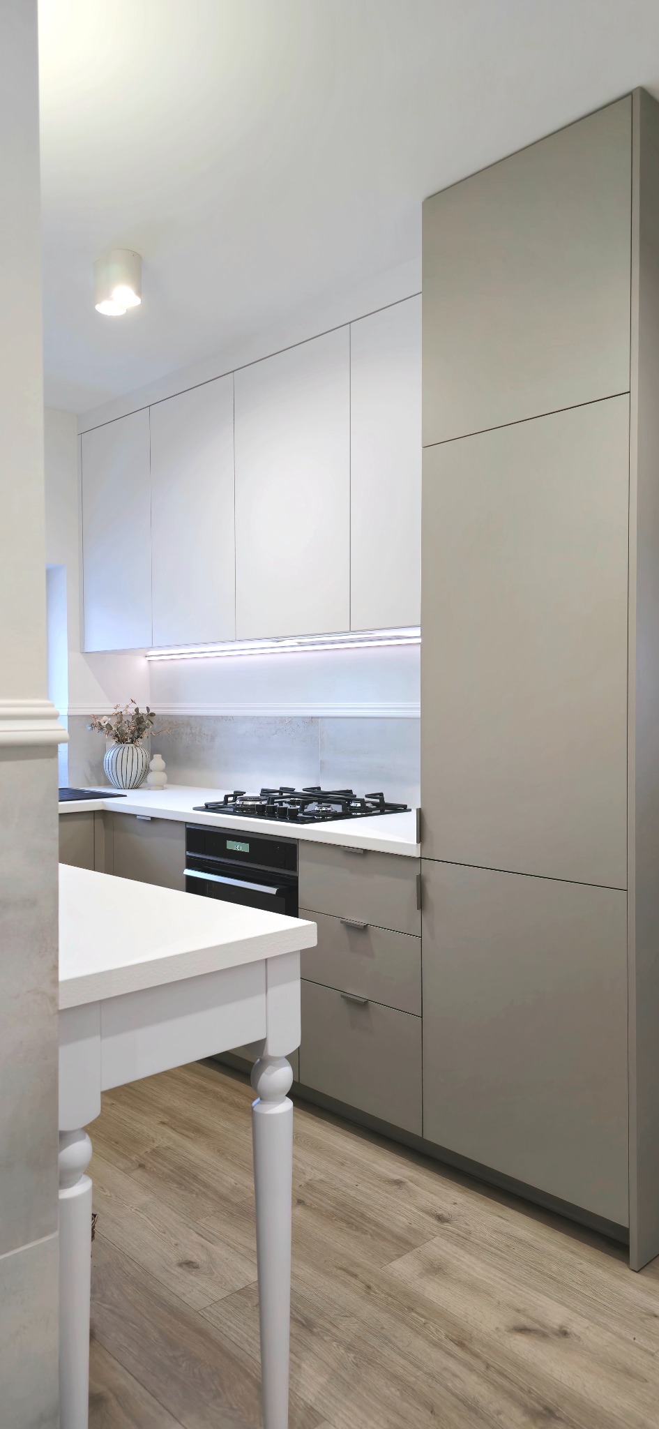 Nowoczesna kuchnia w odcieniach bieli i szarości z drewnianą podłogą, oświetleniem LED pod szafkami i białym stołem z toczonymi nogami na pierwszym planie.