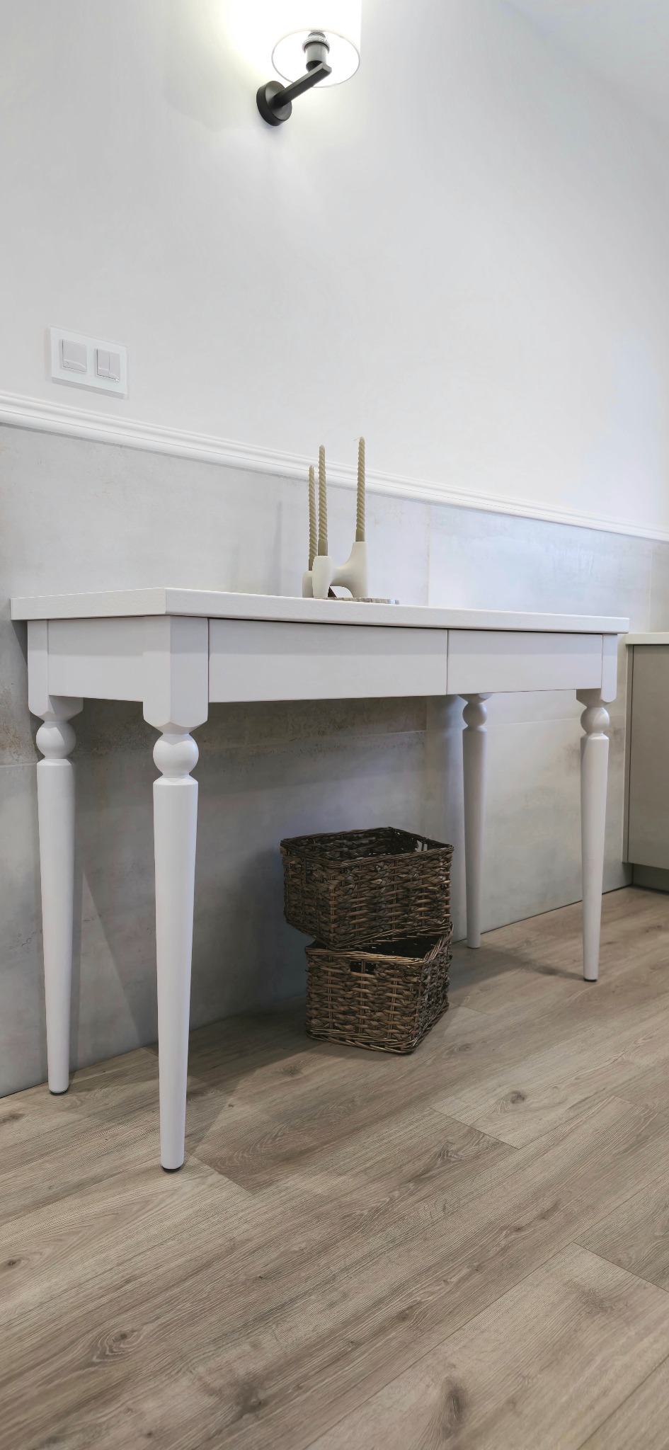 Biały, drewniany stół z toczonymi nogami, ozdobiony świecznikami i ustawiony na podłodze z paneli, z dwoma wiklinowymi koszami umieszczonymi pod blatem, na tle jasnoszarej ściany z listwą...