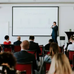 Prelegent w niebieskim garniturze z mikrofonem prezentuje koncepcję 'Lean' na ekranie projektora przed zgromadzoną publicznością podczas szkolenia. Na ekranie widoczne strzałki i napisy 'Jakość...