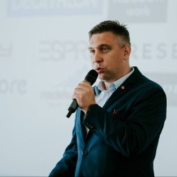 Smart Selling Group Dariusz Gawron - Mężczyzna w granatowym garniturze z mikrofonem, prezentujący na tle tablicy z logotypami firm, podczas szkolenia.