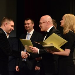 Smart Selling Group Dariusz Gawron - Uroczyste wręczenie certyfikatu lub nagrody, mężczyźni w garniturach ściskają dłonie na tle publiczności.