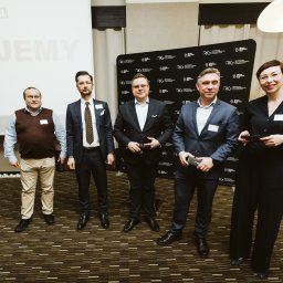 Smart Selling Group Dariusz Gawron - Grupa pięciu osób, w tym czterech mężczyzn w garniturach i jedna kobieta w eleganckim stroju, stojąca na tle ściany z logo RLG w sali konferencyjnej po prezentacji Marshmallow Challenge.