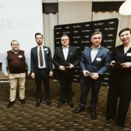 Grupa pięciu osób, w tym czterech mężczyzn w garniturach i jedna kobieta w eleganckim stroju, stojąca na tle ściany z logo RLG w sali konferencyjnej po prezentacji Marshmallow Challenge.