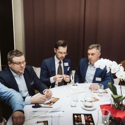 Smart Selling Group Dariusz Gawron - Grupa mężczyzn w garniturach siedzi przy okrągłym stole podczas warsztatów integracyjnych, tworząc konstrukcje z patyczków i papieru, z dekoracjami kwiatowymi i pudełkiem czekoladek.