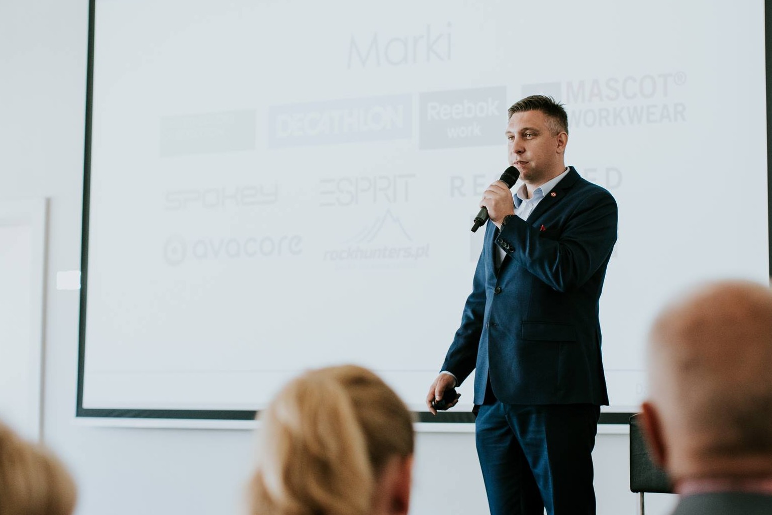 Prelegent w granatowym garniturze przemawia do publiczności z mikrofonem na tle ekranu z logotypami firm.