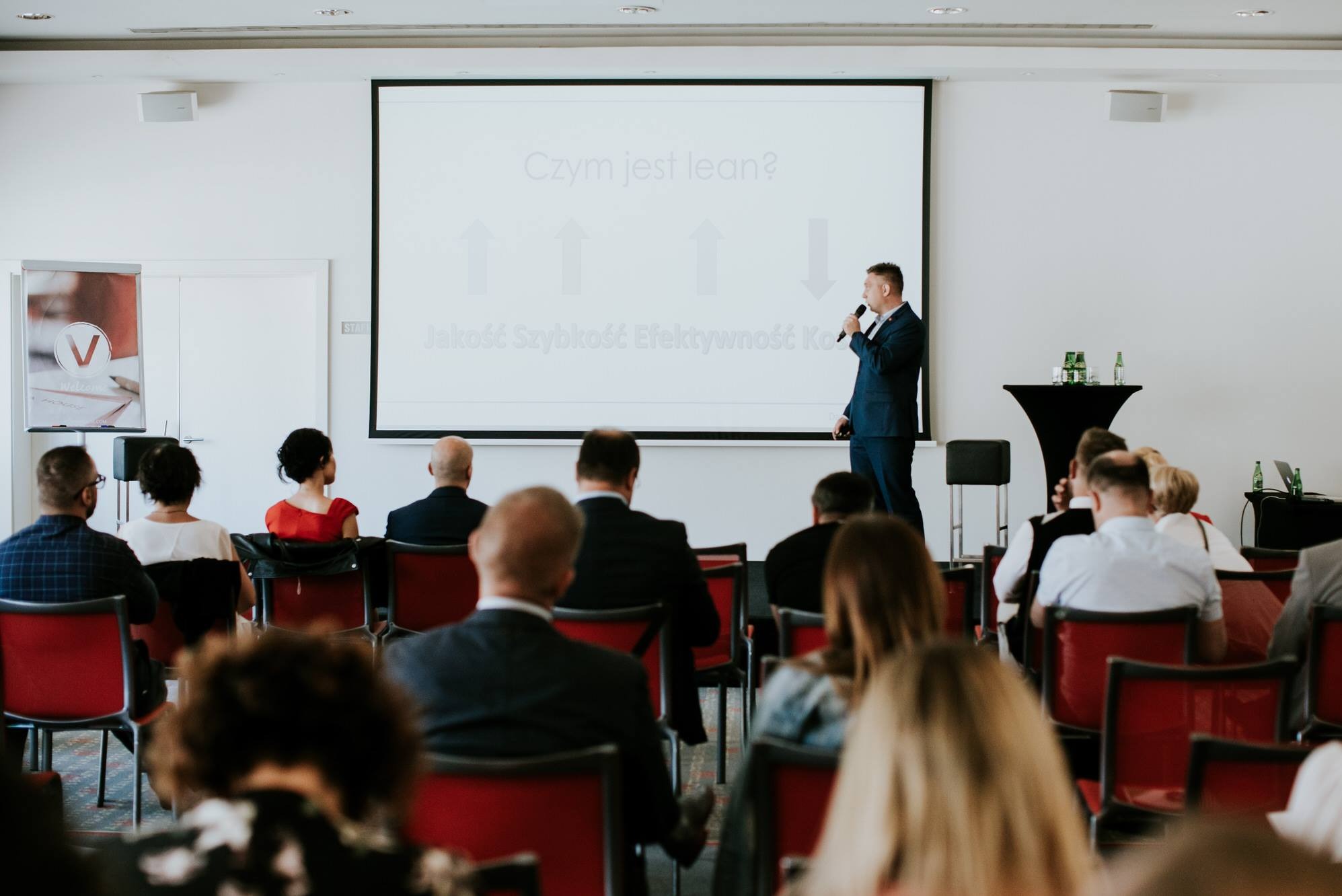 Prelegent w niebieskim garniturze z mikrofonem prezentuje koncepcję 'Lean' na ekranie projektora przed zgromadzoną publicznością podczas szkolenia. Na ekranie widoczne strzałki i napisy 'Jakość...