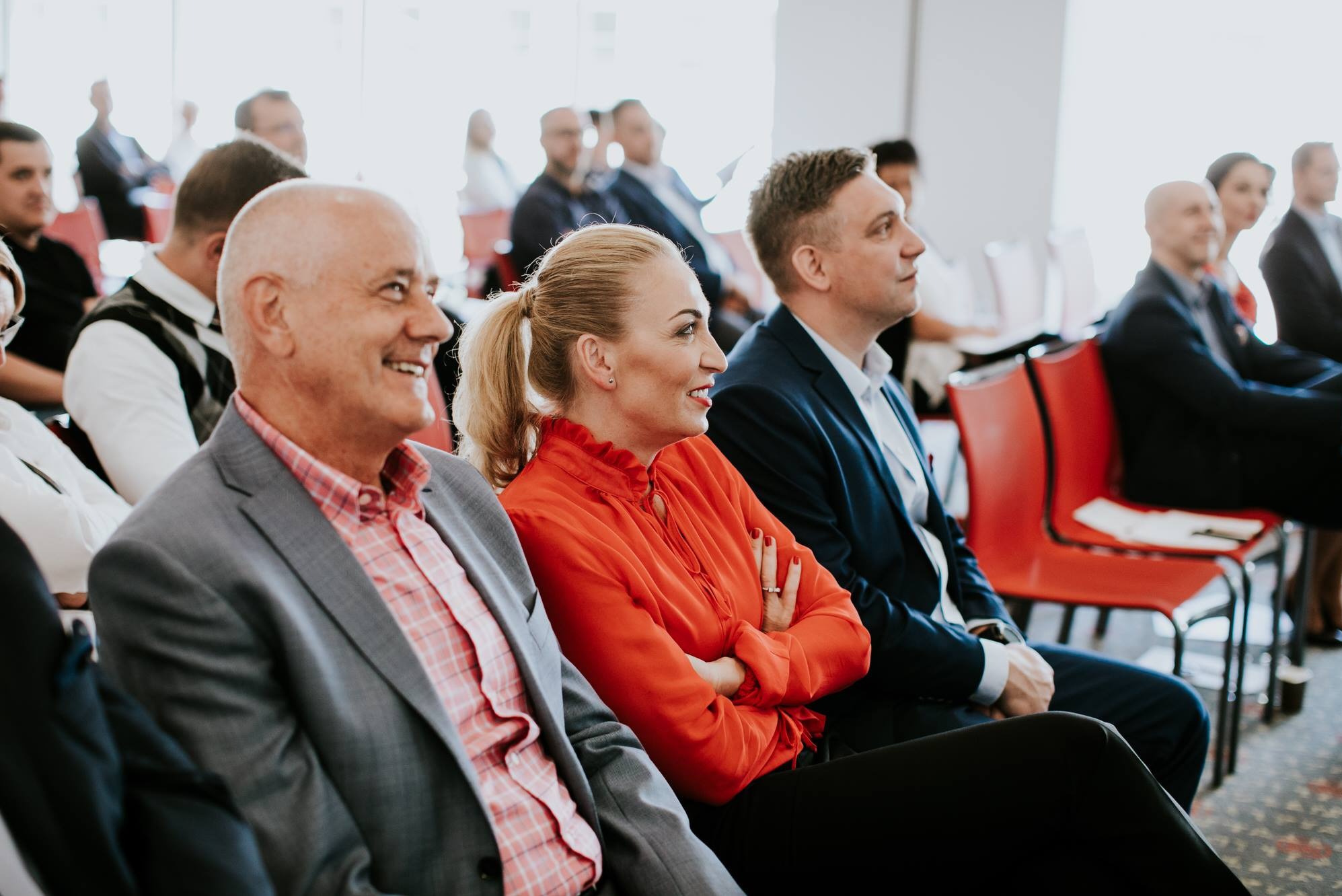 Uczestnicy w rzędach podczas konferencji, mężczyzna w szarej marynarce i różowej koszuli w kratę uśmiecha się, kobieta w pomarańczowej bluzce z falbanami ma skrzyżowane ręce, mężczyzna w granatowym...