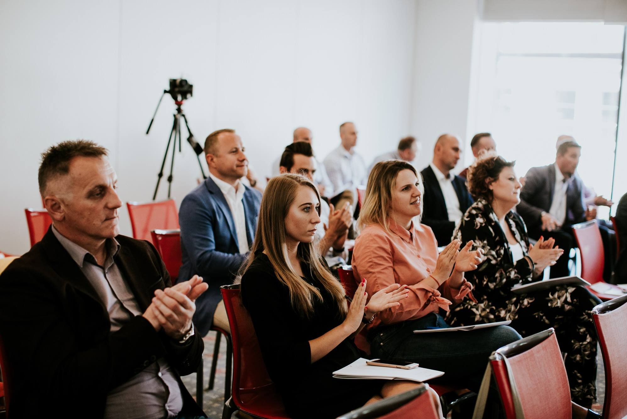 Grupa osób siedzi w rzędach na czerwonych krzesłach, klaszcząc podczas prezentacji w jasnym pomieszczeniu; w tle widoczny statyw z kamerą.