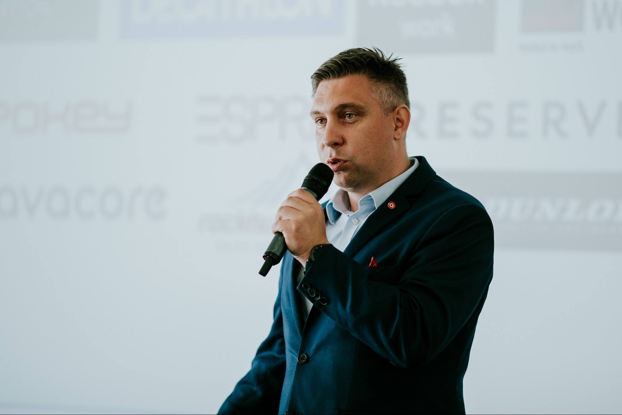 Mężczyzna w granatowym garniturze z mikrofonem, prezentujący na tle tablicy z logotypami firm, podczas szkolenia.