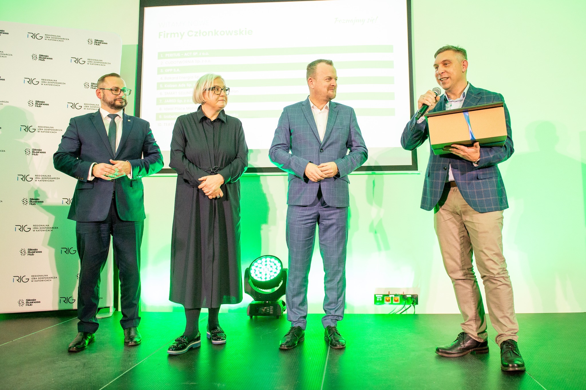 Prezentacja dla firm członkowskich Regionalnej Izby Gospodarczej: mężczyzna w garniturze wręcza nagrodę w pudełku podczas oficjalnego spotkania na scenie.
