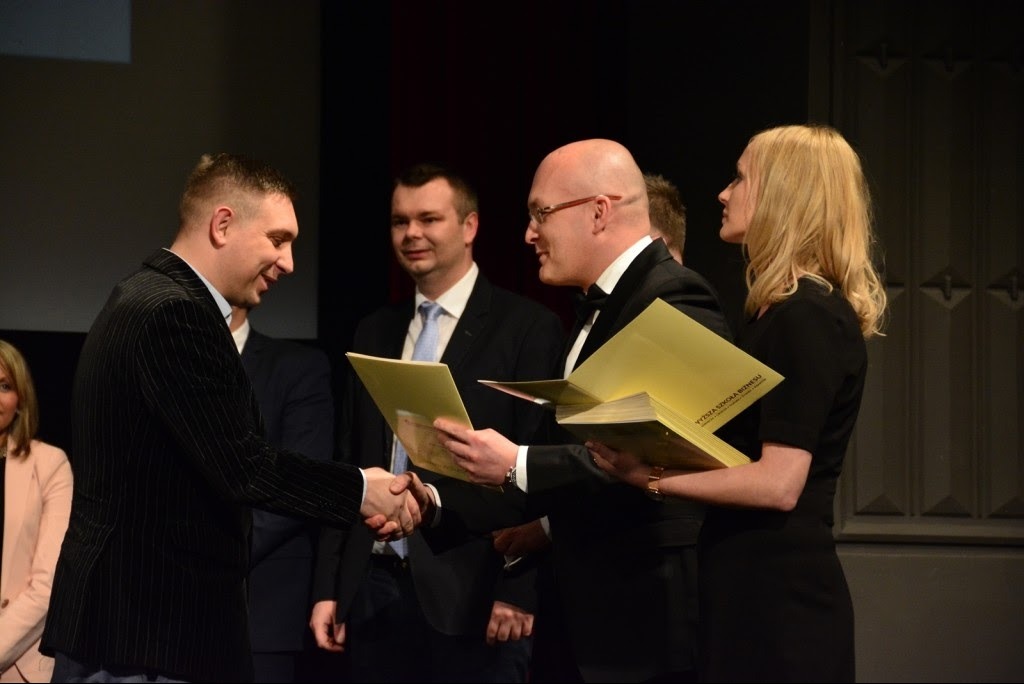 Uroczyste wręczenie certyfikatu lub nagrody, mężczyźni w garniturach ściskają dłonie na tle publiczności.