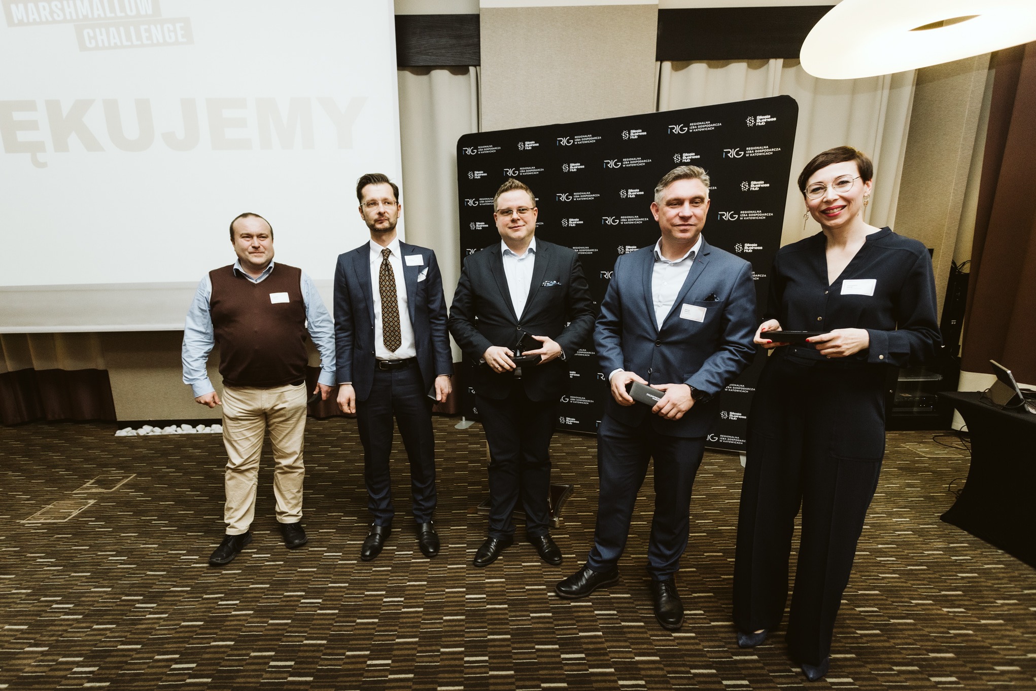 Grupa pięciu osób, w tym czterech mężczyzn w garniturach i jedna kobieta w eleganckim stroju, stojąca na tle ściany z logo RLG w sali konferencyjnej po prezentacji Marshmallow Challenge.