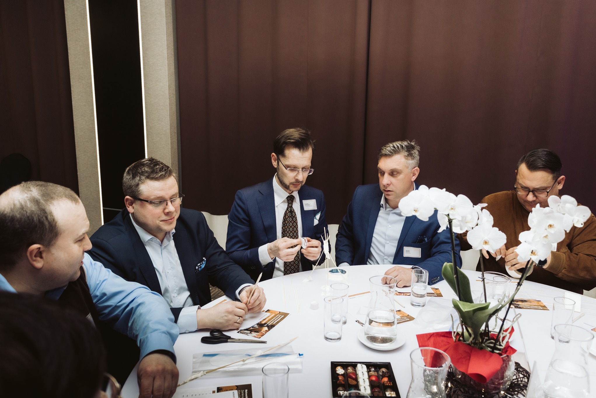 Grupa mężczyzn w garniturach siedzi przy okrągłym stole podczas warsztatów integracyjnych, tworząc konstrukcje z patyczków i papieru, z dekoracjami kwiatowymi i pudełkiem czekoladek.