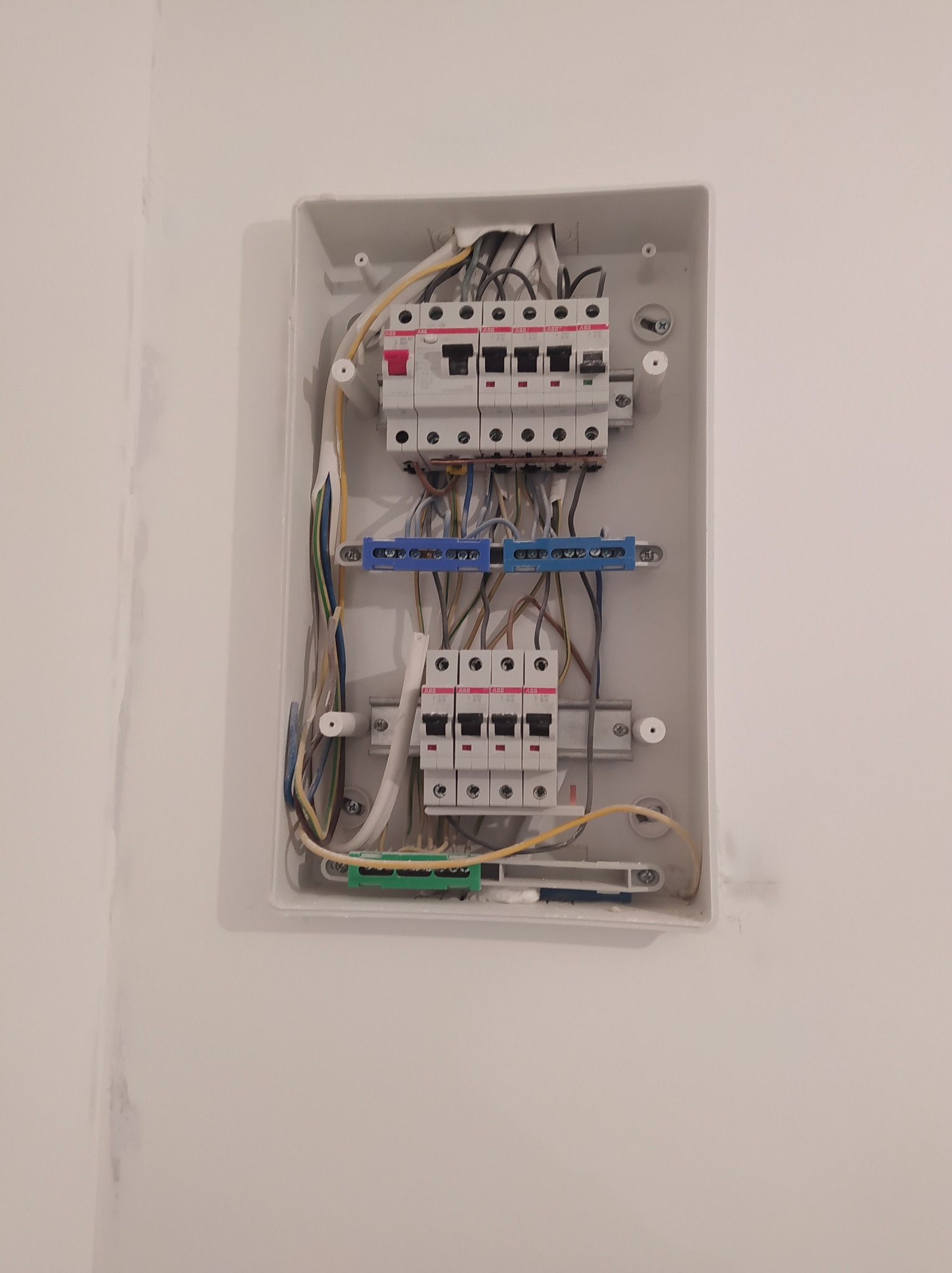 Otwarta, biała skrzynka elektryczna z zainstalowanymi bezpiecznikami, listwami zaciskowymi i okablowaniem na białej ścianie. Widoczne ślady po malowaniu na ścianie w rogu.