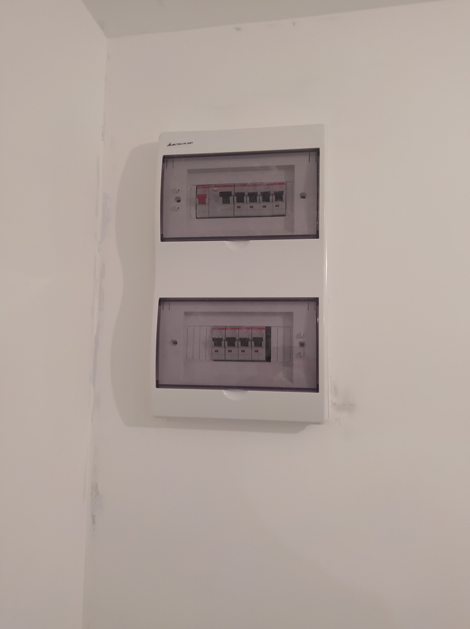 Dwa białe rozdzielnice elektryczne z widocznymi bezpiecznikami zamontowane na jasnej ścianie, widoczne ślady po malowaniu na rogu ściany.