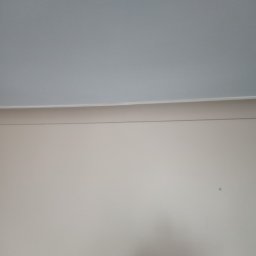 Lukas - Świeżo pomalowana ściana w odcieniach beżu z białym sufitem, widoczny cień i mała plamka na ścianie.