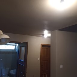 Lukas - Wnętrze po odświeżeniu ścian z widocznym wejściem do łazienki i drewnianymi drzwiami do innego pomieszczenia, oświetlone lampami sufitowymi.