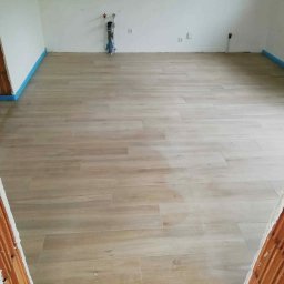 Lukas - Jasne, drewnopodobne panele ułożone w pustym pomieszczeniu z fragmentami surowych ścian. Niebieska taśma zabezpieczająca przy podłodze. Widoczne instalacje na ścianie.